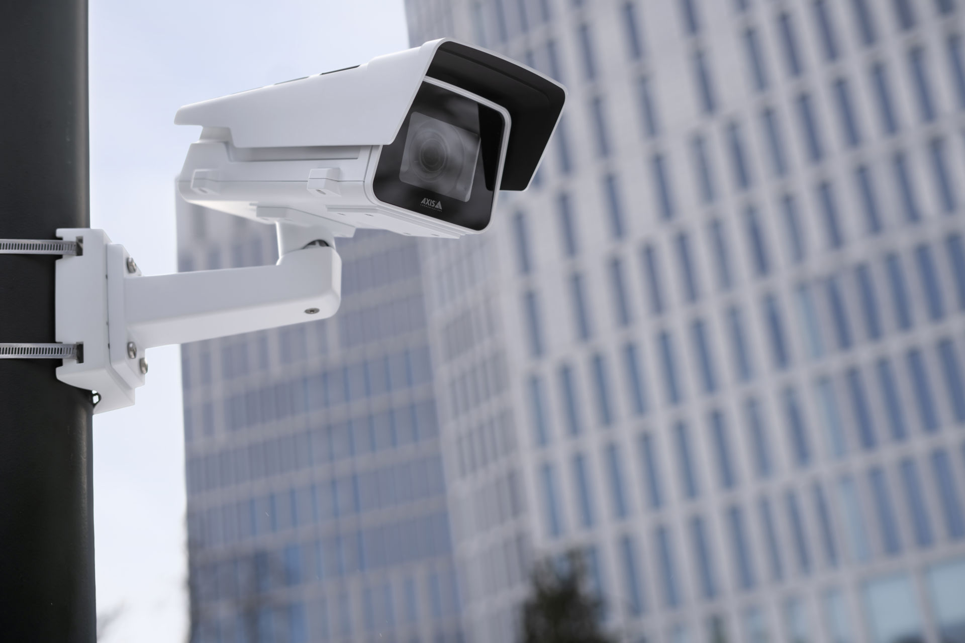 A white AXIS P1385-E box camera with a sunshield mounted on a black pole, overlooking a modern glass office building.