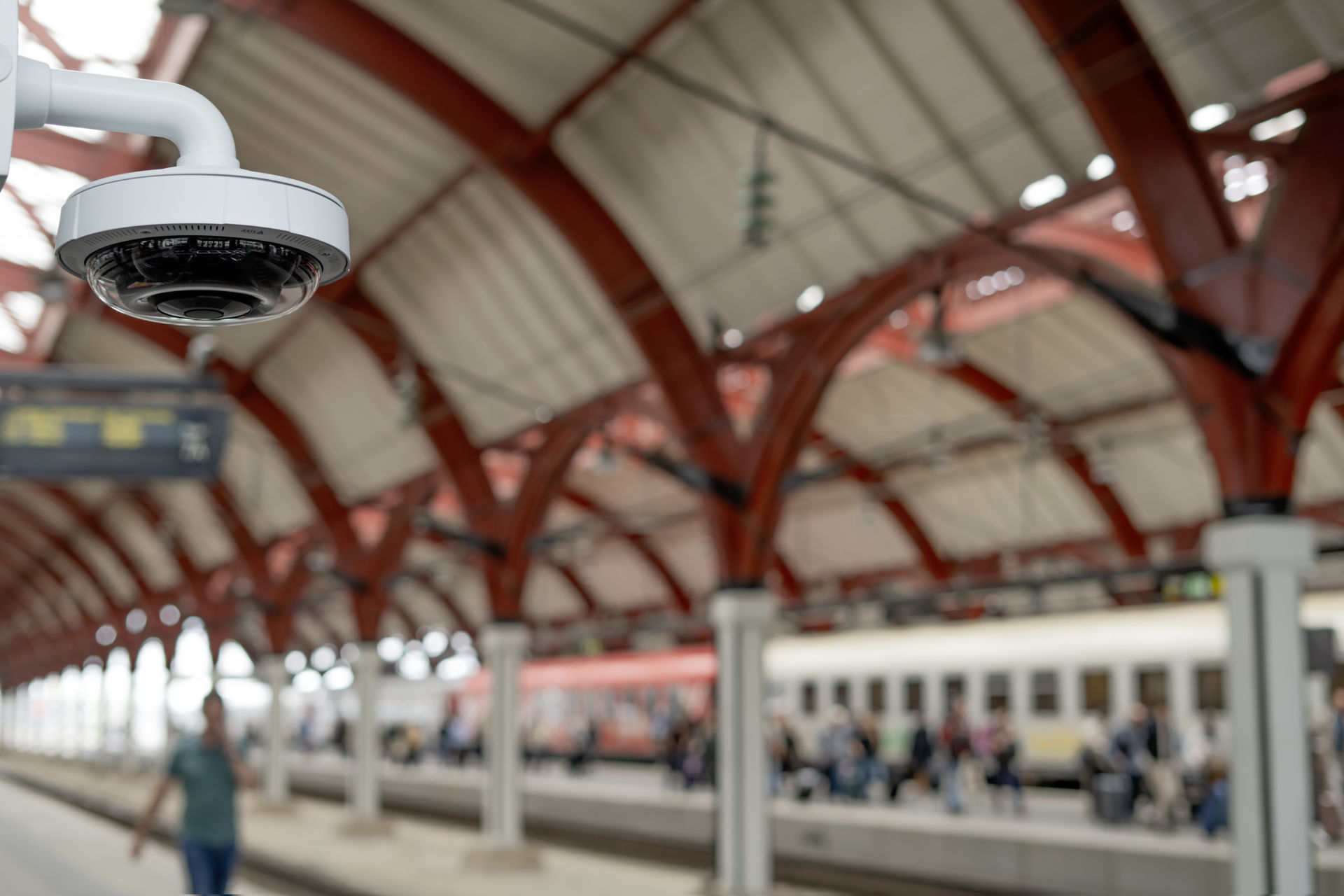 AXIS P3735-PLE multidirectional camera mounted on a white pendant arm overlooking a busy train station platform with red arches.