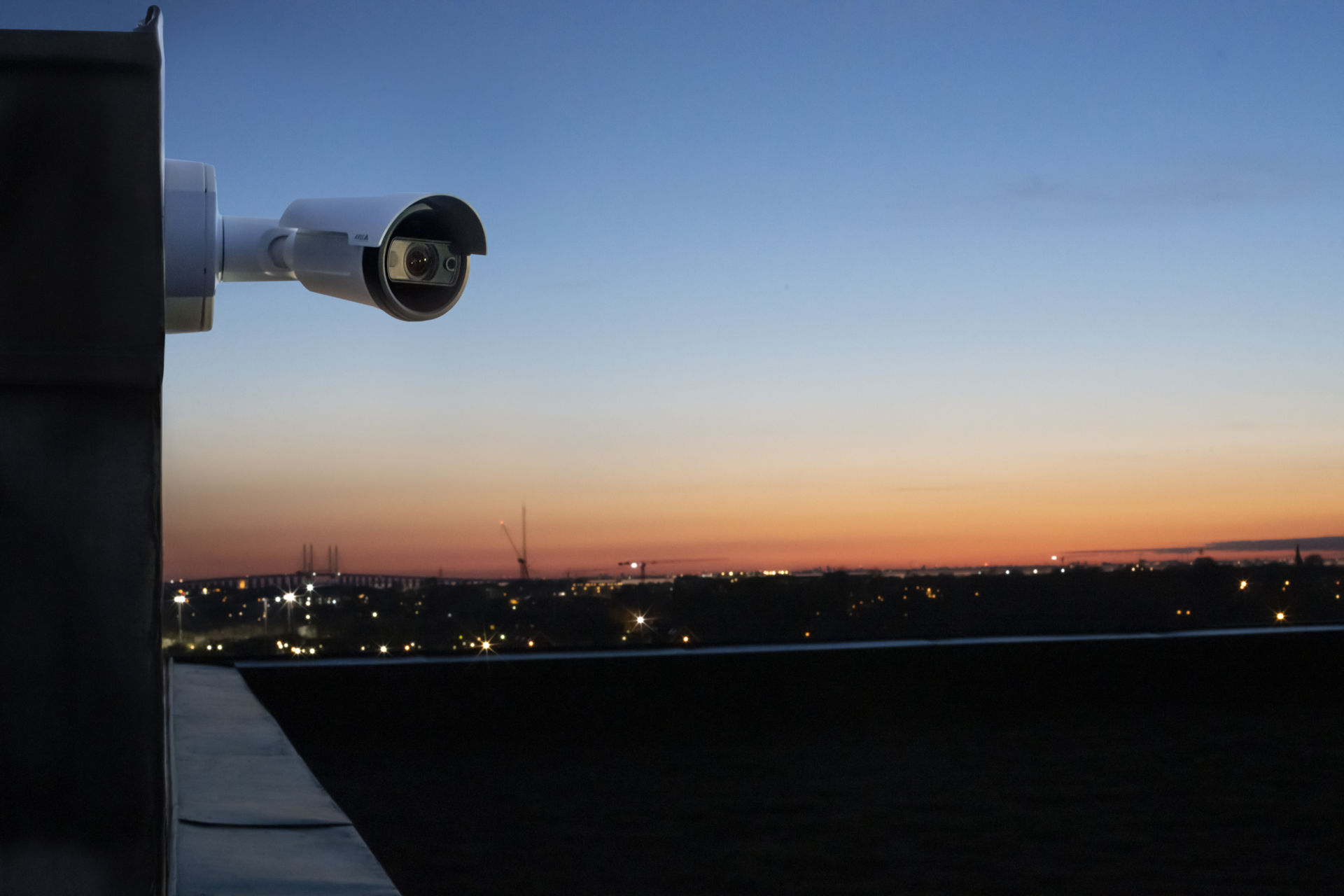 A white AXIS bullet security camera mounted on a rooftop ledge, overlooking a city skyline during a deep orange and blue sunset.