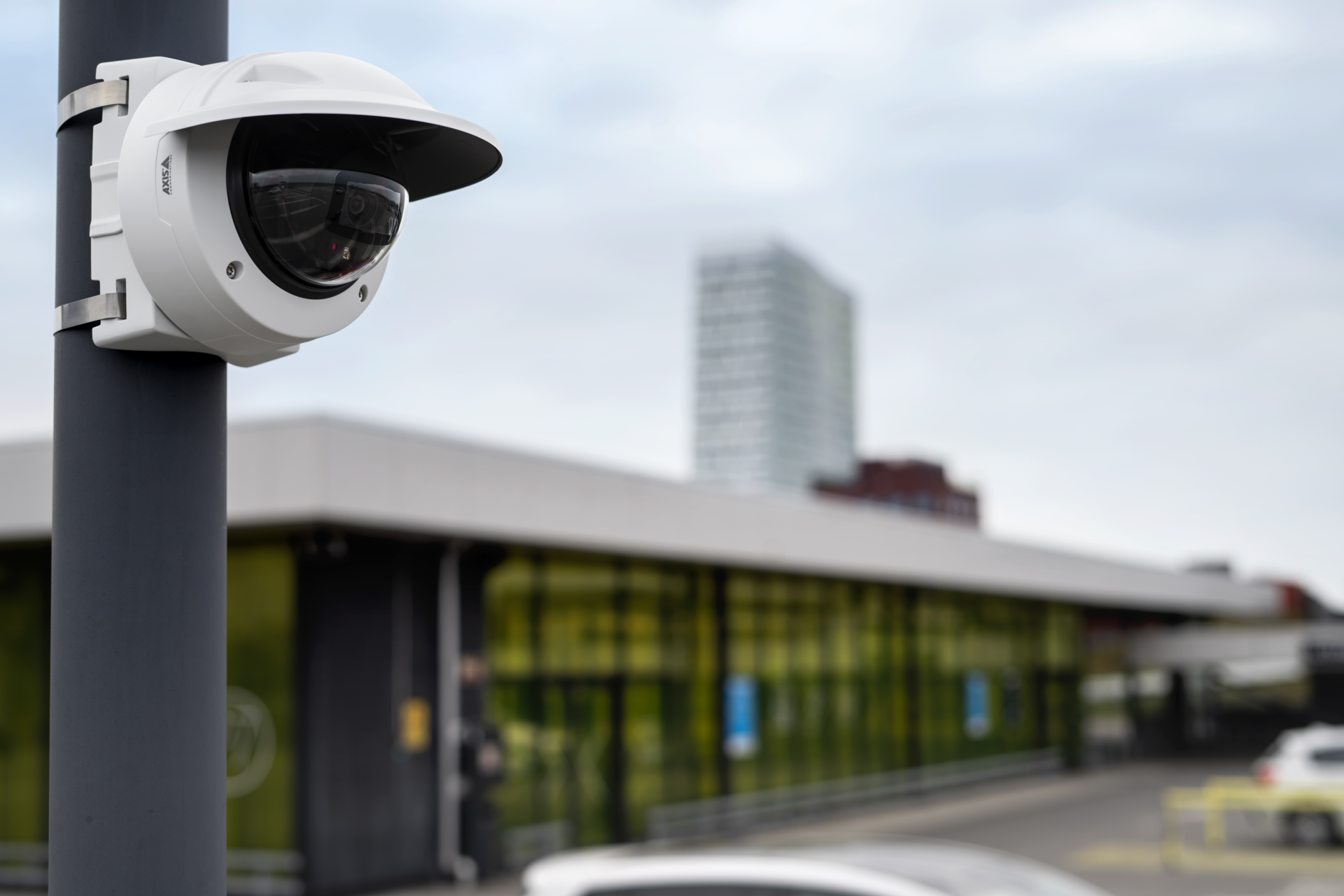 A white AXIS dome security camera with a weather shield mounted to a grey pole overlooking a parking area and modern building.