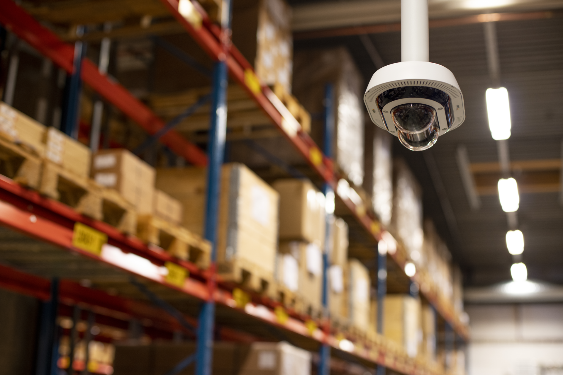 A white AXIS dual-sensor multidirectional dome camera mounted to a ceiling pipe in a large warehouse. The background shows blurred industrial shelving units stacked with cardboard boxes and bright overhead lights.