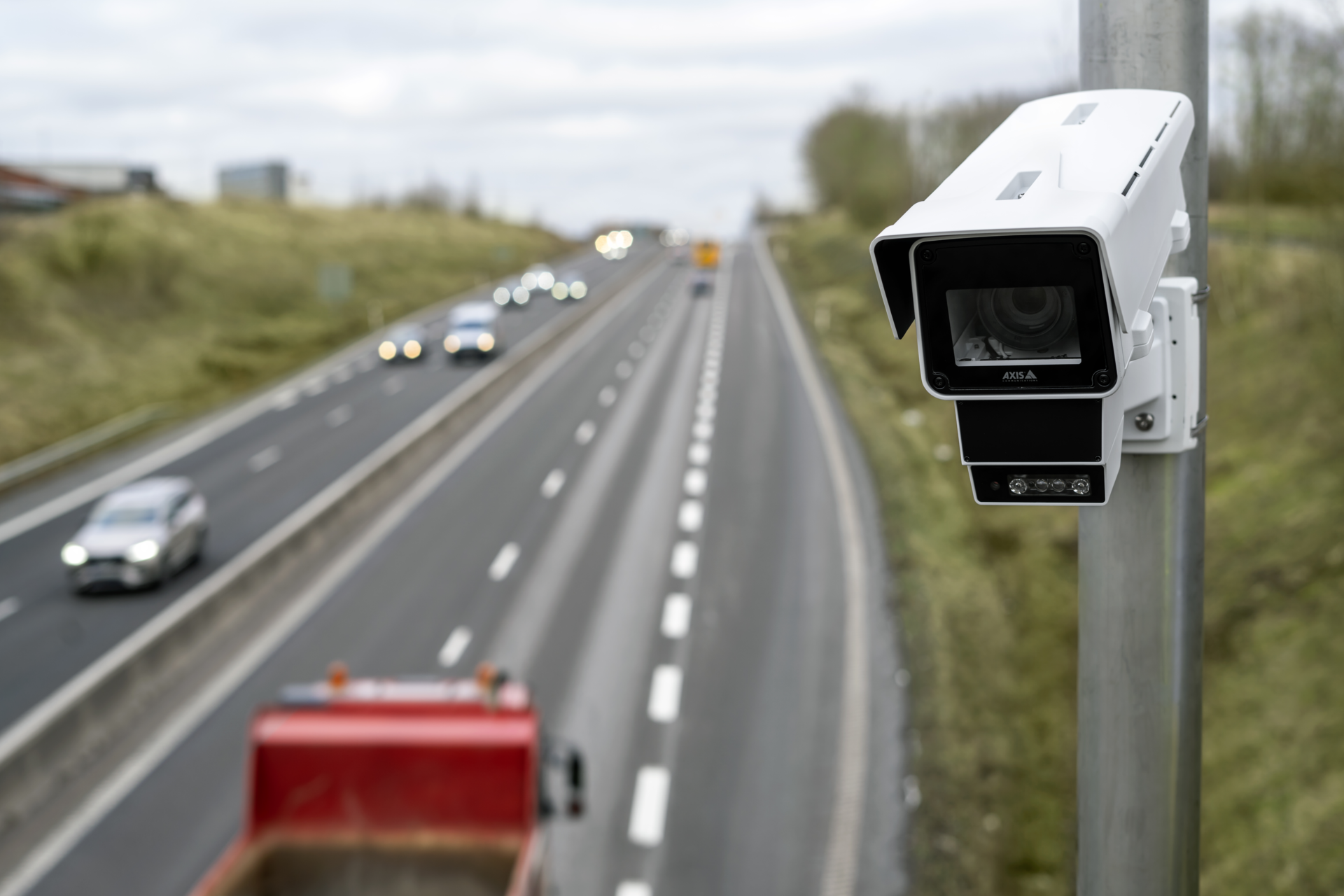 AXIS Q1686-DLE radar-video fusion camera mounted on a metal pole, monitoring daytime vehicle traffic and a red truck on a multi-lane highway.