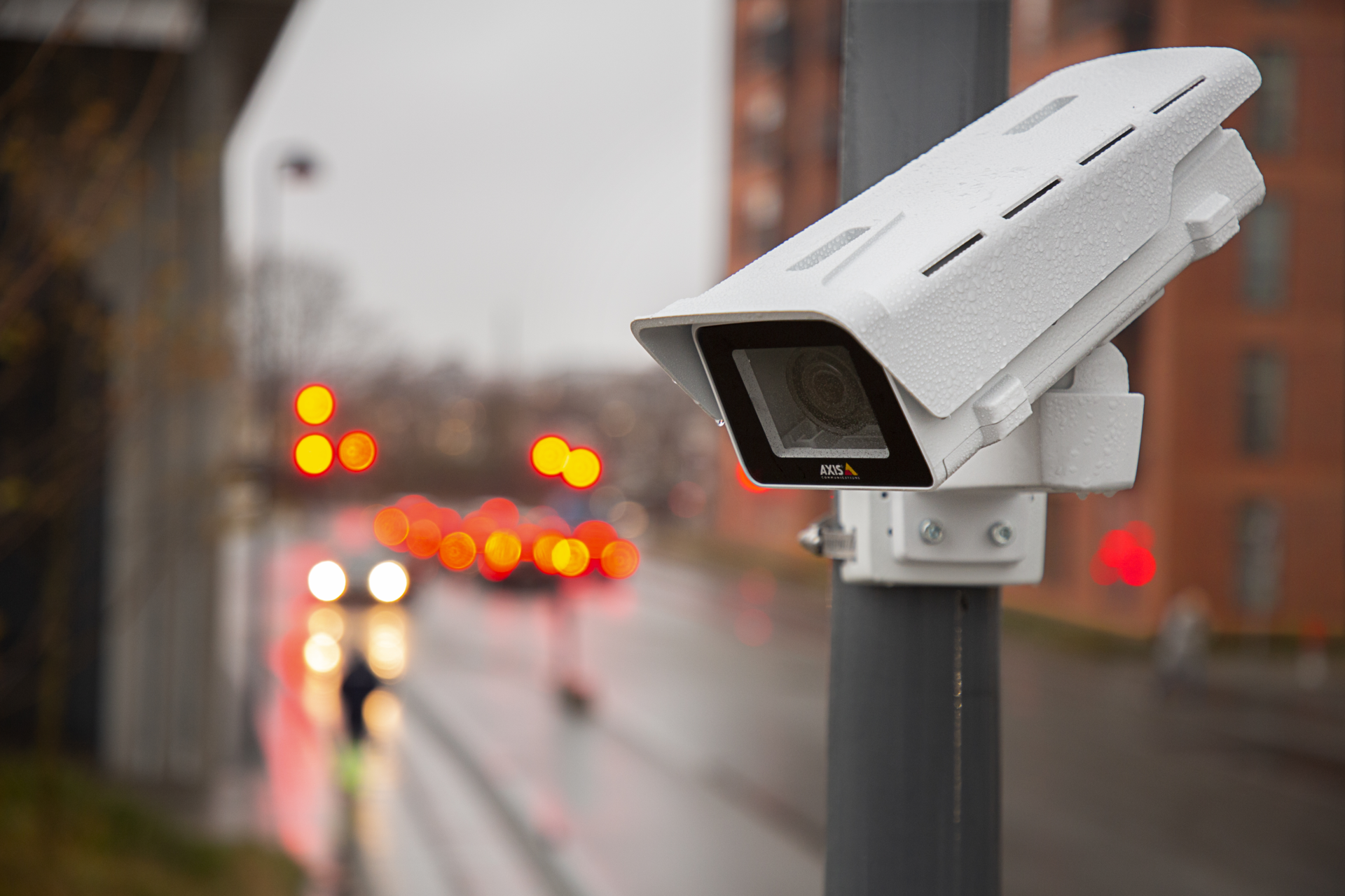 AXIS M1137-E Network Camera mounted on a street pole, with blurred traffic lights and vehicles visible in the background.