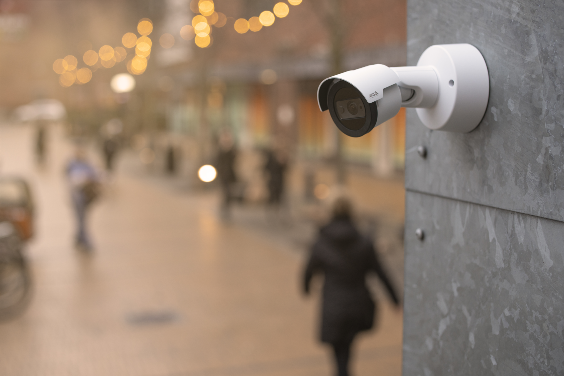 An AXIS M2036-LE bullet network camera mounted on a grey wall, overlooking a blurred outdoor pedestrian shopping street with warm string lights.