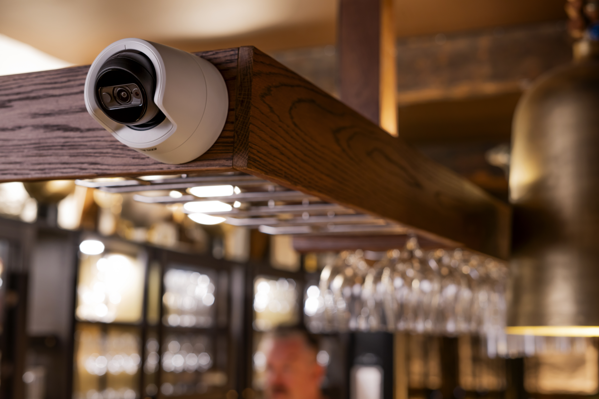 AXIS M3125-LVE Network Camera mounted on a wooden ceiling beam in a restaurant bar area, with wine glasses suspended below and warm ambient lighting in the background.