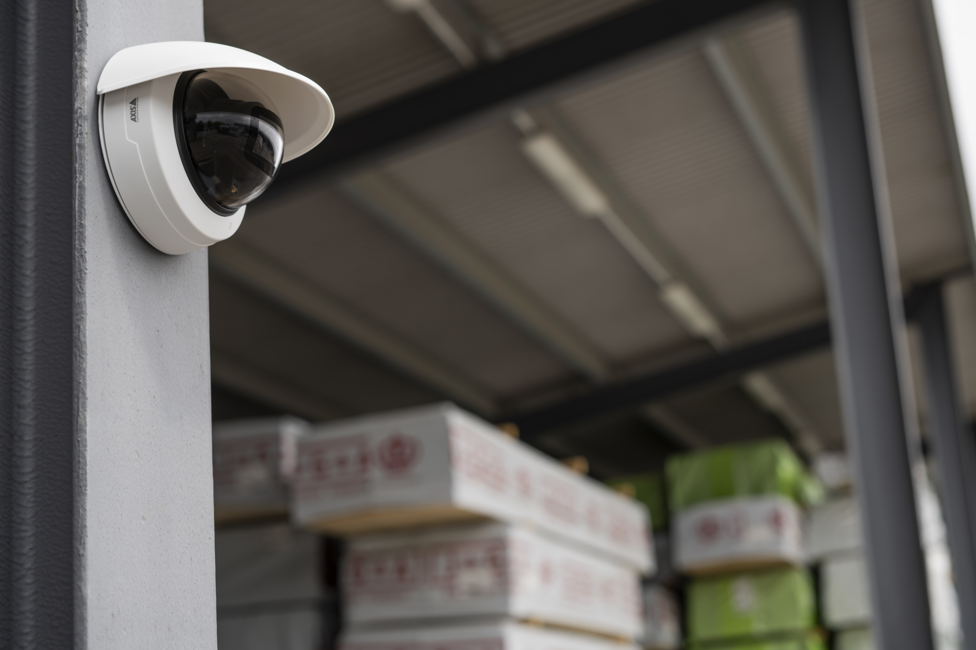 AXIS M4225-LVE-W dome camera mounted on a concrete pillar at the entrance of a covered outdoor storage facility, with stacked boxes visible on shelving in the background.
