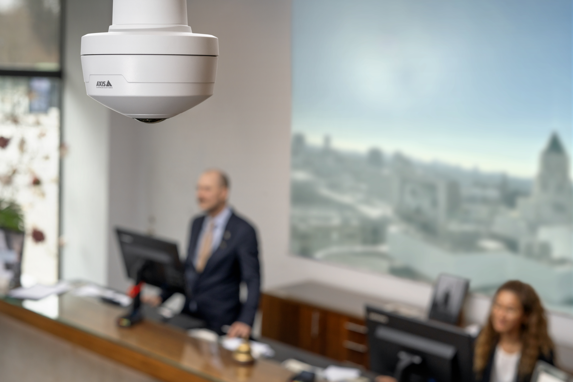 AXIS M4327-P dome camera mounted on a hotel reception ceiling, with staff members visible at the front desk in the background.