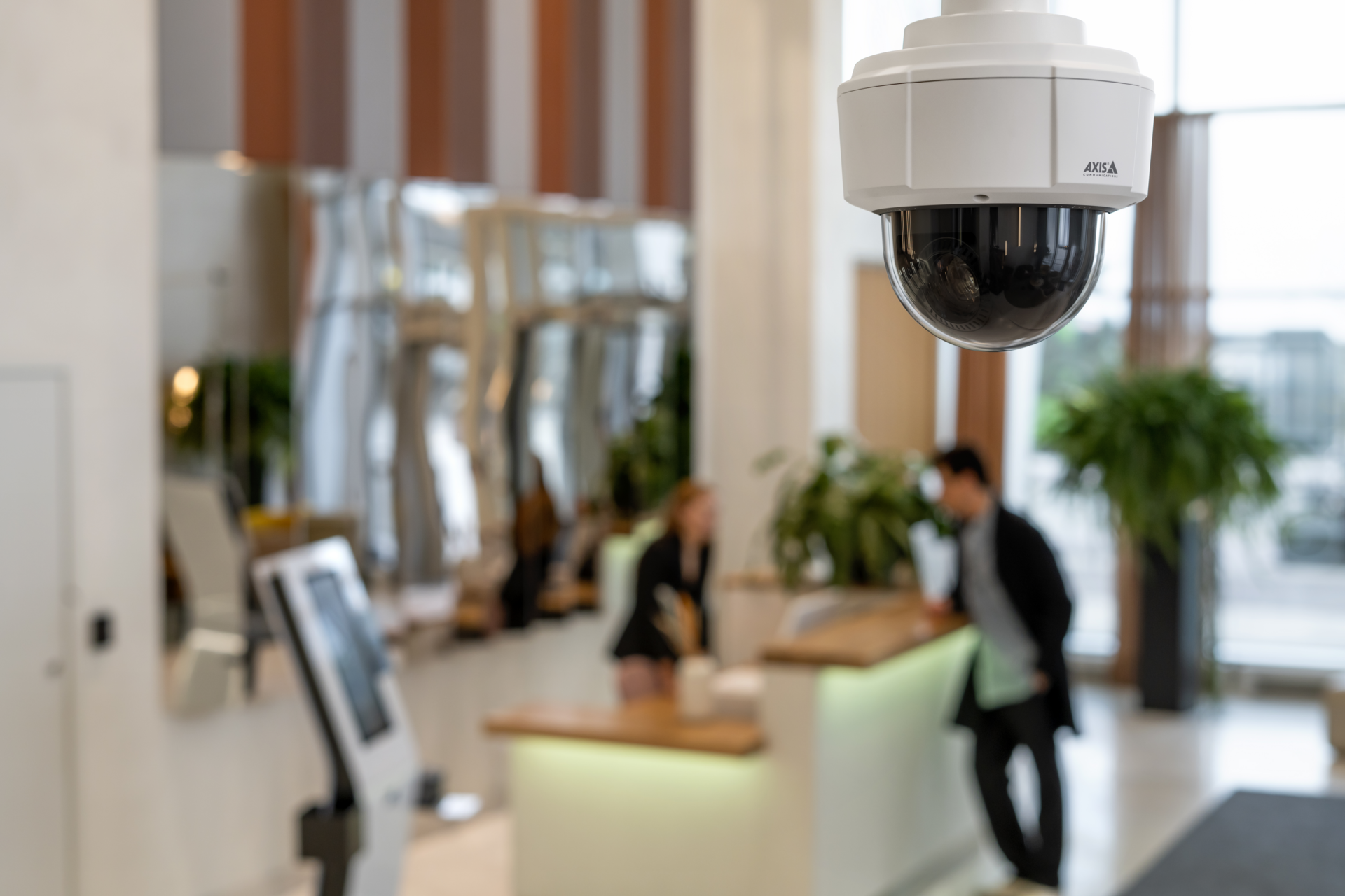 AXIS M5526-E dome PTZ camera mounted on a ceiling, overlooking a modern office reception area where a receptionist and a visitor are visible in the background.
