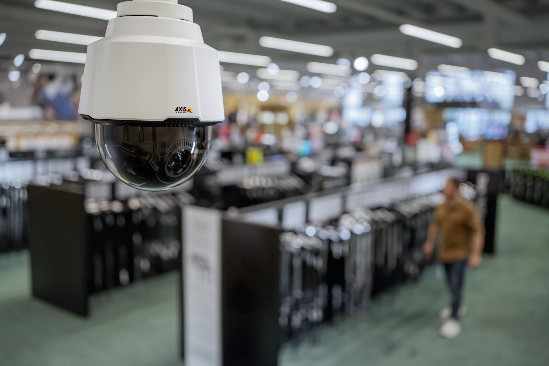 AXIS P5654-E Mk II PTZ dome camera mounted from a retail store ceiling, with product display aisles and a shopper visible in the background.