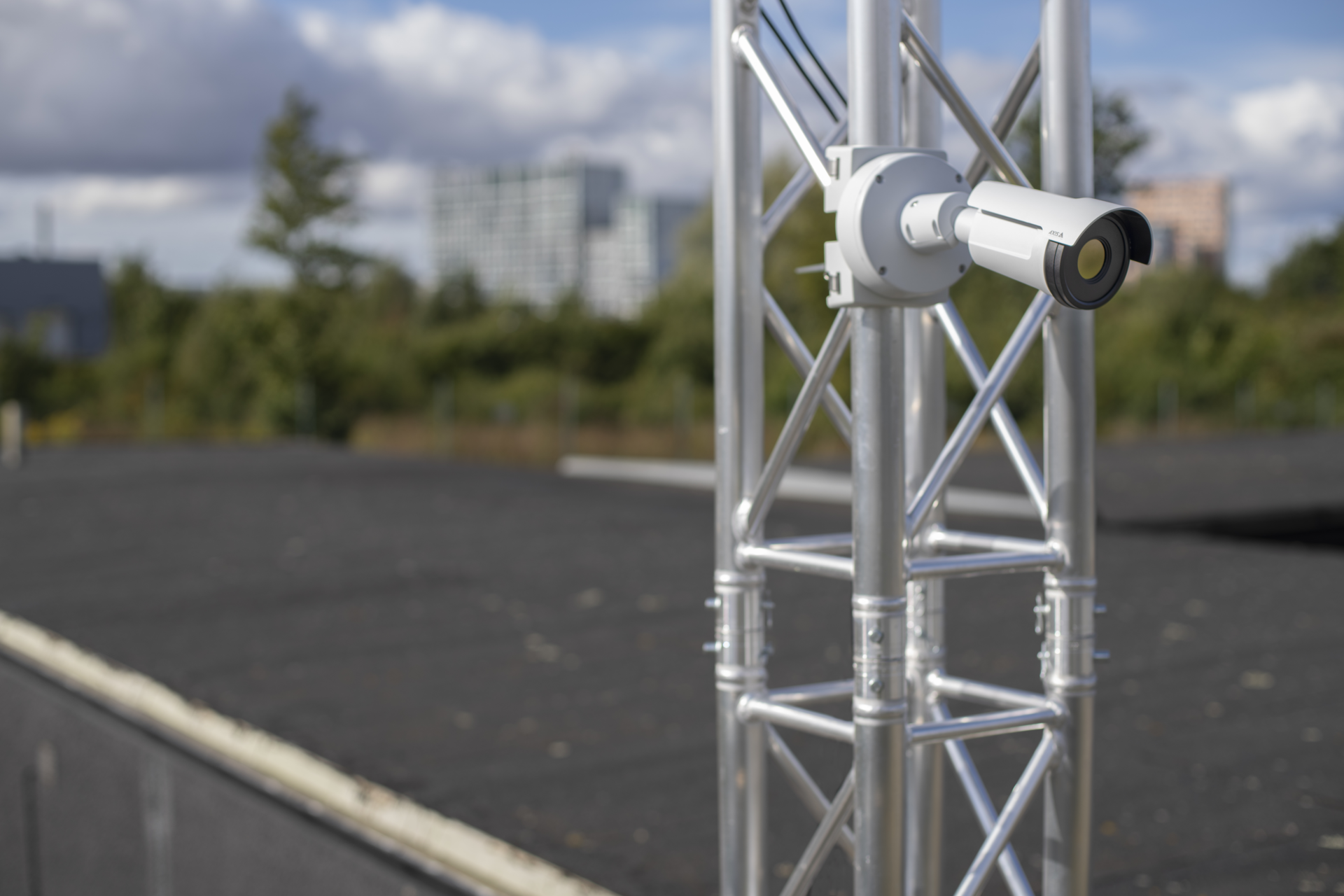 AXIS Q1961-TE thermal camera mounted on a galvanised steel truss structure outdoors, overlooking a rooftop area with trees and buildings in the background.