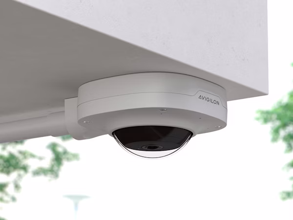 A white Avigilon Alta H5A 360 camera mounted to an outdoor ceiling soffit with a side-entry conduit. The dome-shaped security camera is positioned to provide a panoramic view of the surrounding greenery.