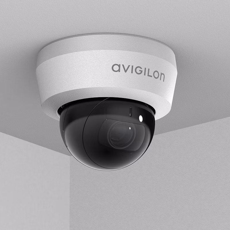 An Avigilon H6 Mini Dome camera mounted on a white ceiling in a corner, featuring a compact white housing and a dark tinted dome lens.