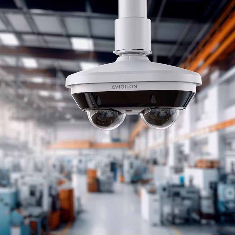 A white Avigilon H6A Dual Head security camera with two adjustable lenses mounted to a ceiling pipe in a bright, blurred industrial factory setting.
