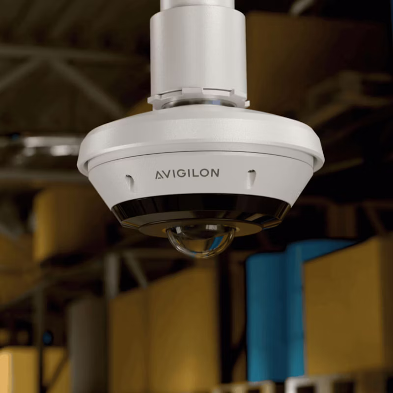 Close-up of a white Avigilon H6A Fisheye camera mounted on a pendant ceiling pipe in a warehouse setting with blurred storage crates in the background.