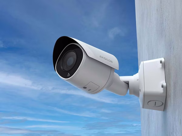 A white Avigilon H6SL bullet security camera mounted on a concrete wall against a clear blue sky with light clouds.