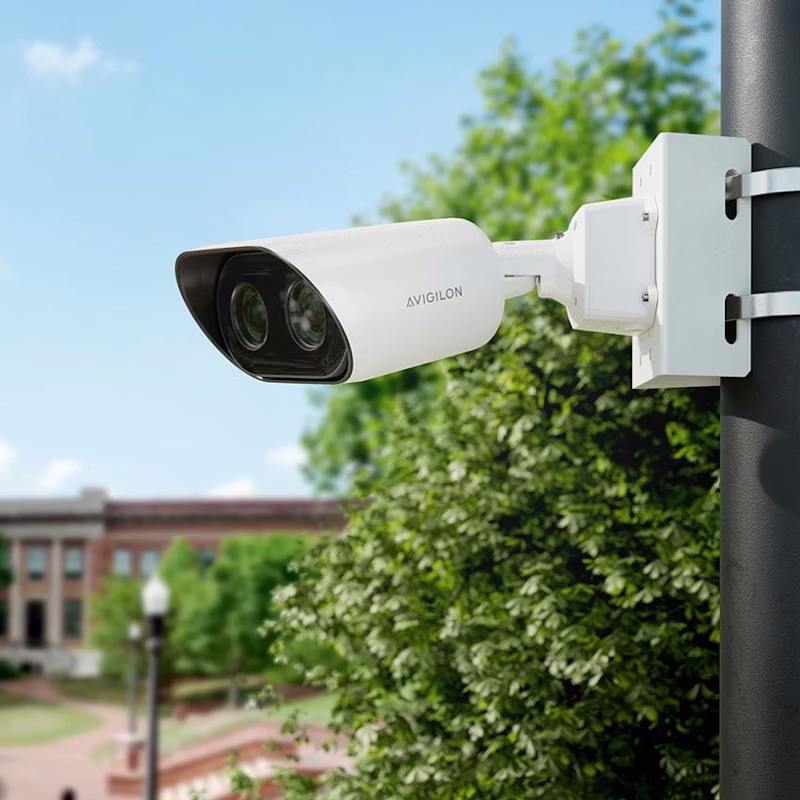An Avigilon L6A Enterprise LPR camera mounted on a black street pole with a green park and brick building in the blurred background.