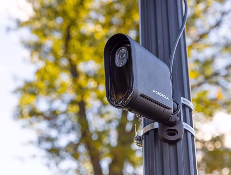 An Avigilon L6Q quick-deploy security camera mounted to a fluted metal street pole using metal bands, with a blurred background of autumn trees.
