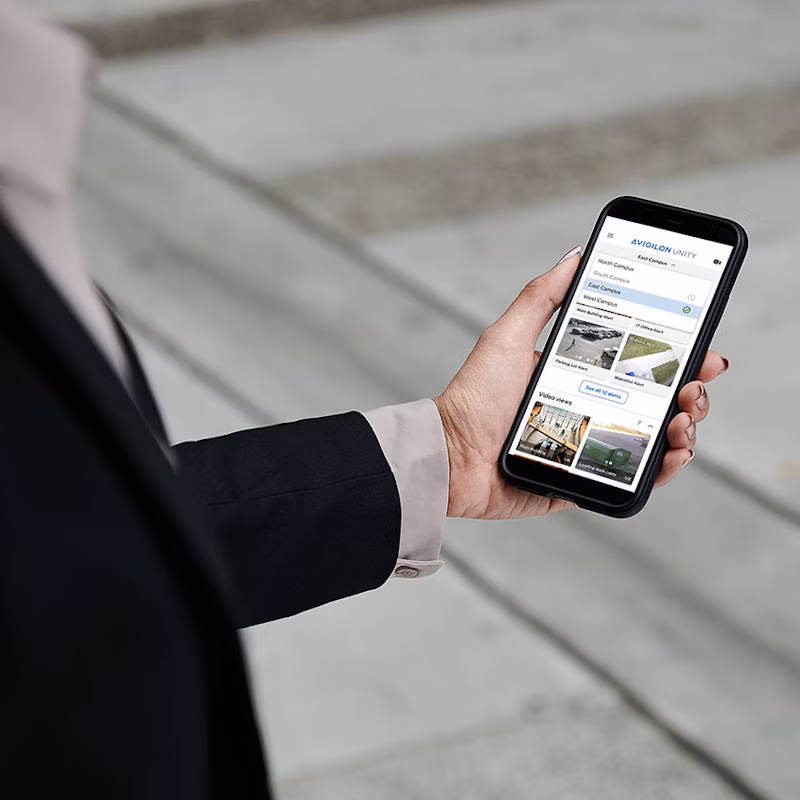 A person in a black blazer holding a smartphone displaying the Avigilon Unity mobile app interface, showing campus alerts and live video feeds.
