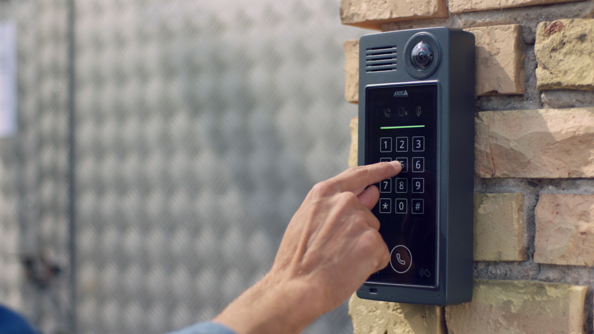 A close-up of a person’s hand entering a code on the illuminated keypad of an AXIS A8207-VE Mk II Network Video Door Station mounted on a light-colored brick wall.