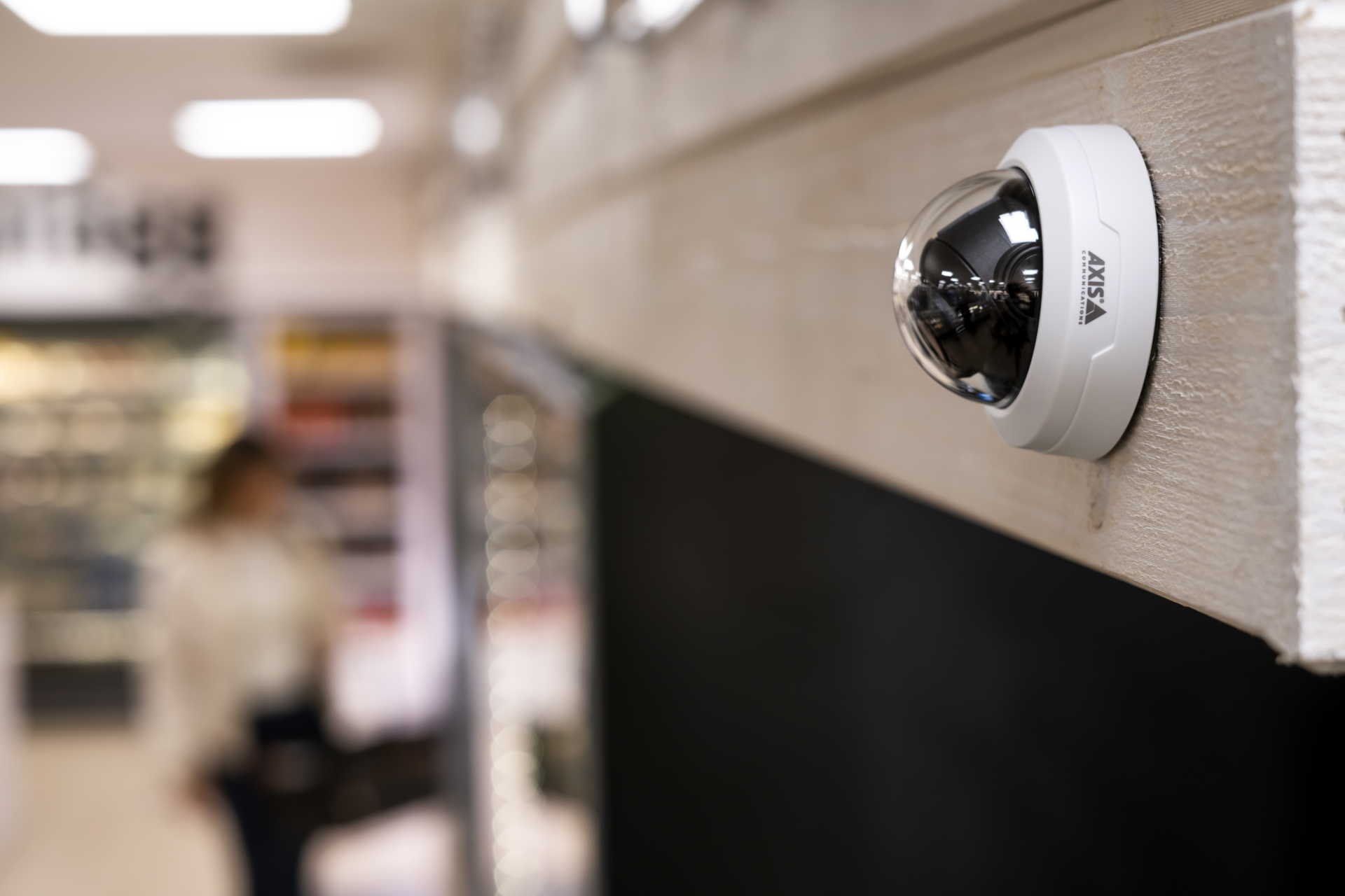 An AXIS F4108 dome sensor unit mounted on a light wood beam in a retail store, with a blurred shopper and grocery aisles in the background.