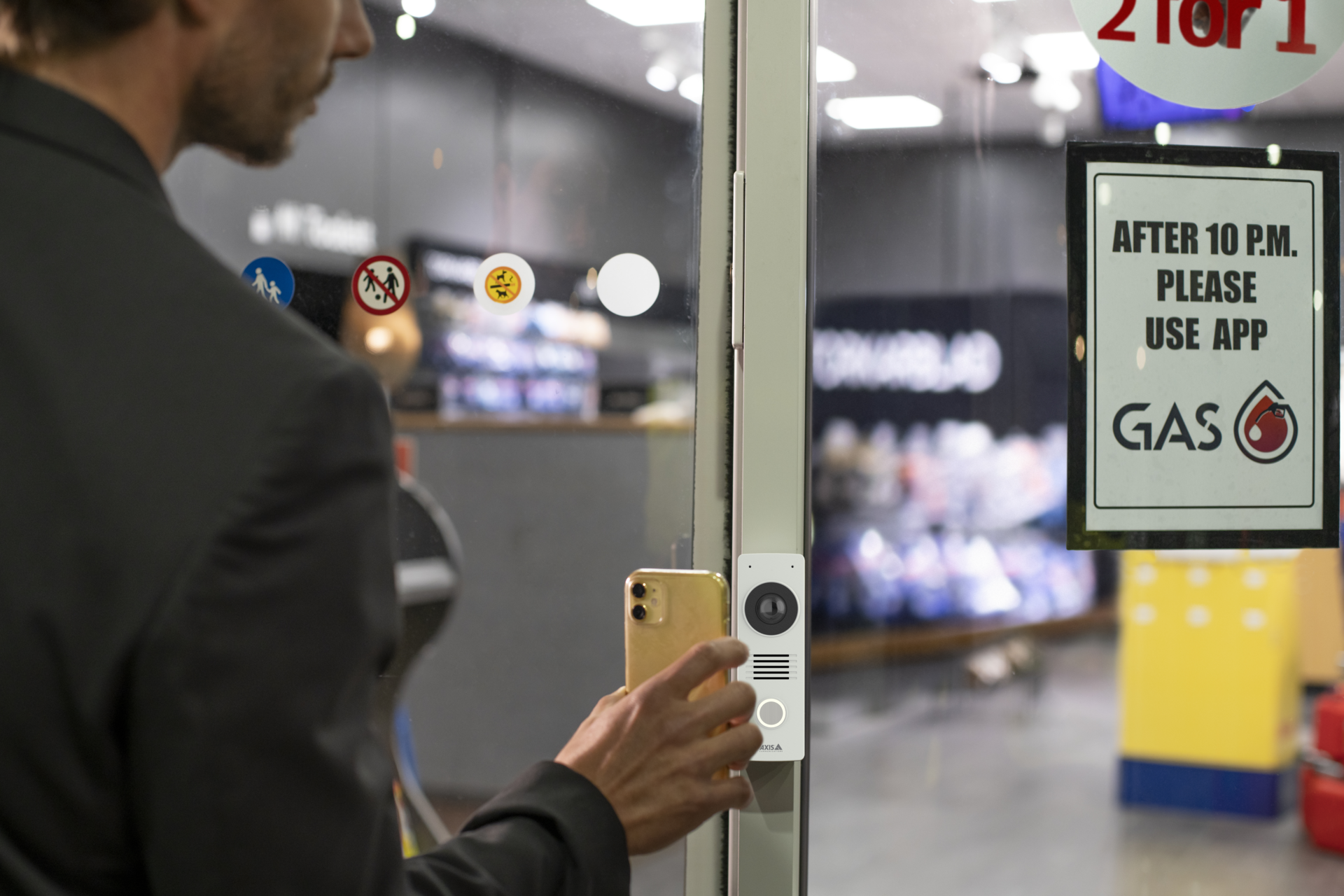 A man in a suit holding a smartphone up to an AXIS I8116-E Network Video Intercom mounted on a glass door at a service station.