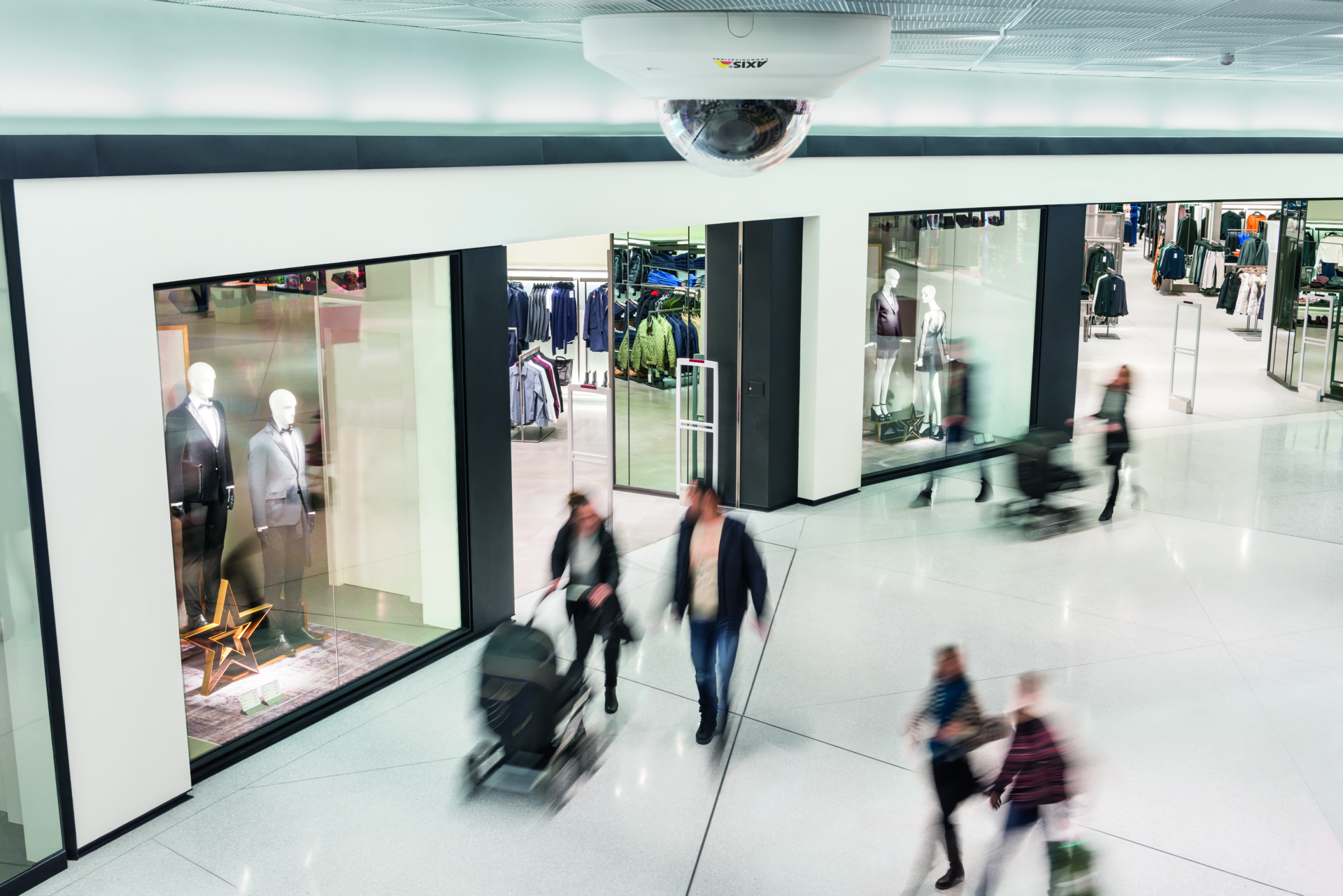 A white AXIS dome security camera mounted on a high ceiling overlooking a busy, modern shopping mall corridor with blurred shoppers walking past high-end clothing store displays
