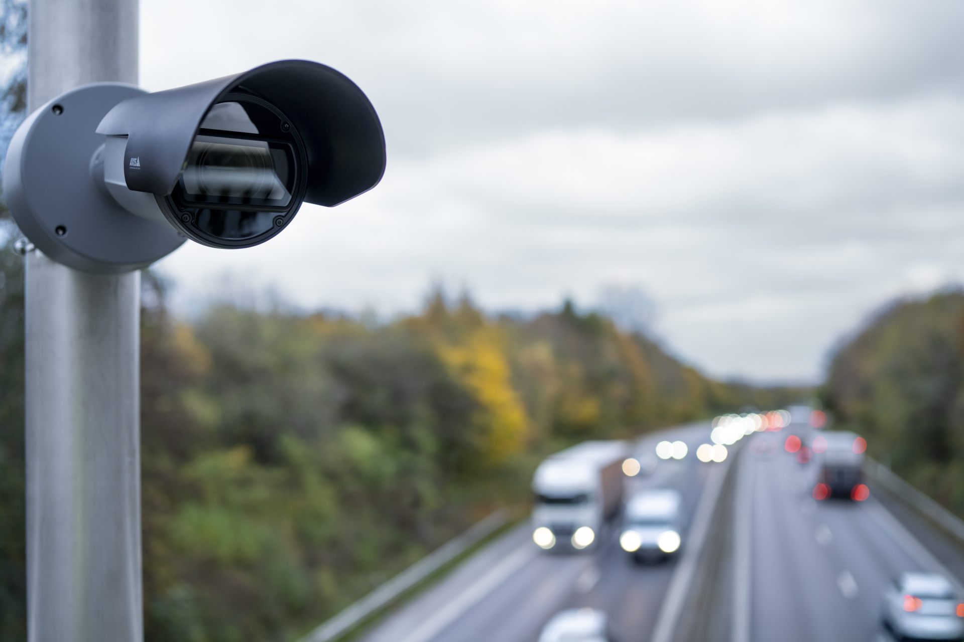 A close-up of an AXIS Q1806-LE License Plate Verifier camera mounted on a metal pole, overlooking a blurred multi-lane motorway with moving vehicles under an overcast sky.