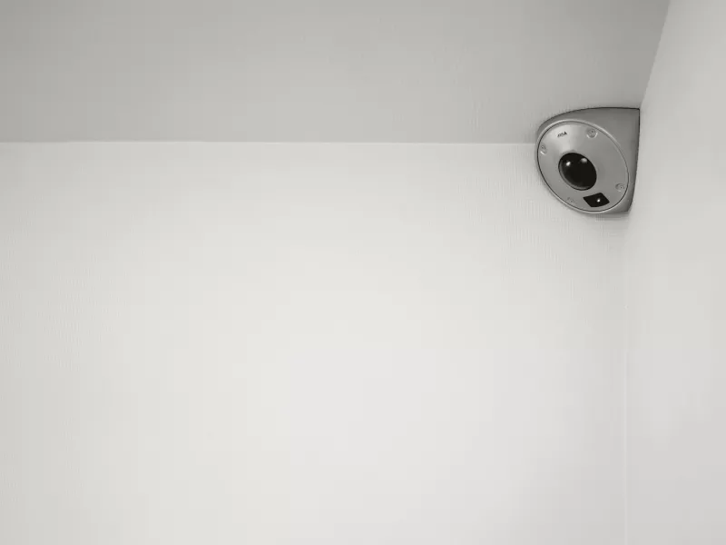 A high-angle, black and white shot of an AXIS Q9227-SLV stainless steel corner-mount security camera installed at the intersection of two white walls and a ceiling.