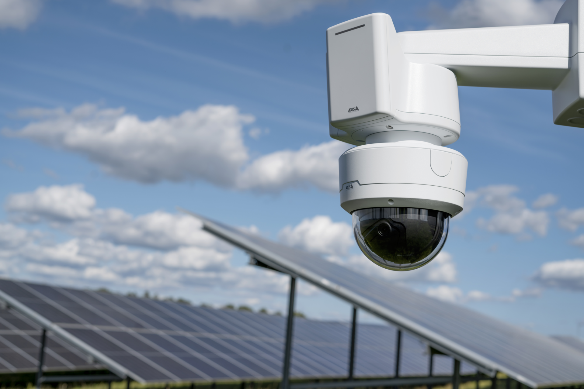 An AXIS Q3839-PVE outdoor PTZ dome security camera mounted on a wall bracket, positioned in front of rows of solar panels under a partly cloudy blue sky.