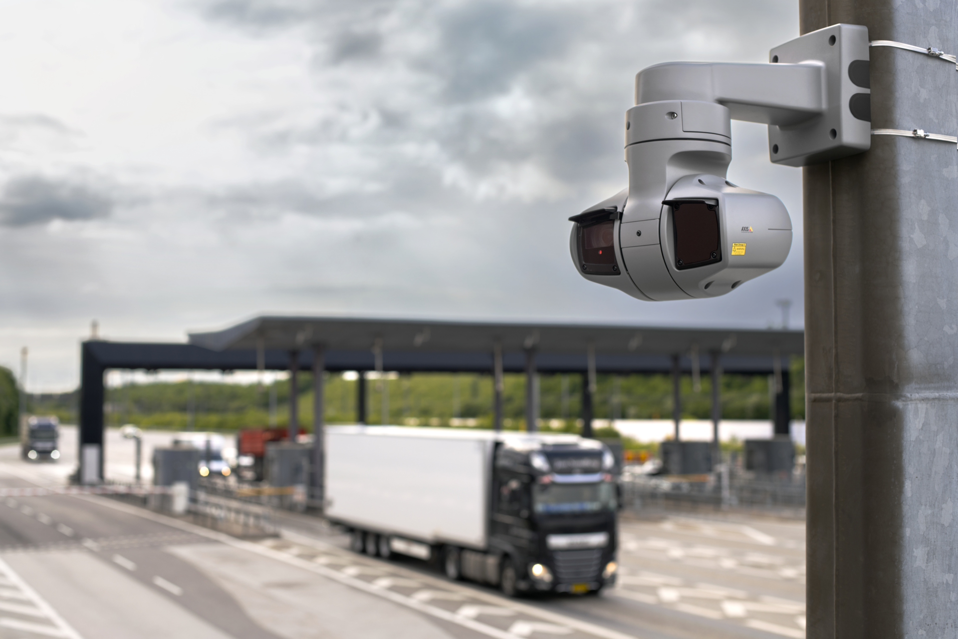 AXIS Q6225-LE PTZ network camera mounted on a pole overlooking a busy highway toll station, with a large articulated truck and multiple vehicles approaching the toll booths.