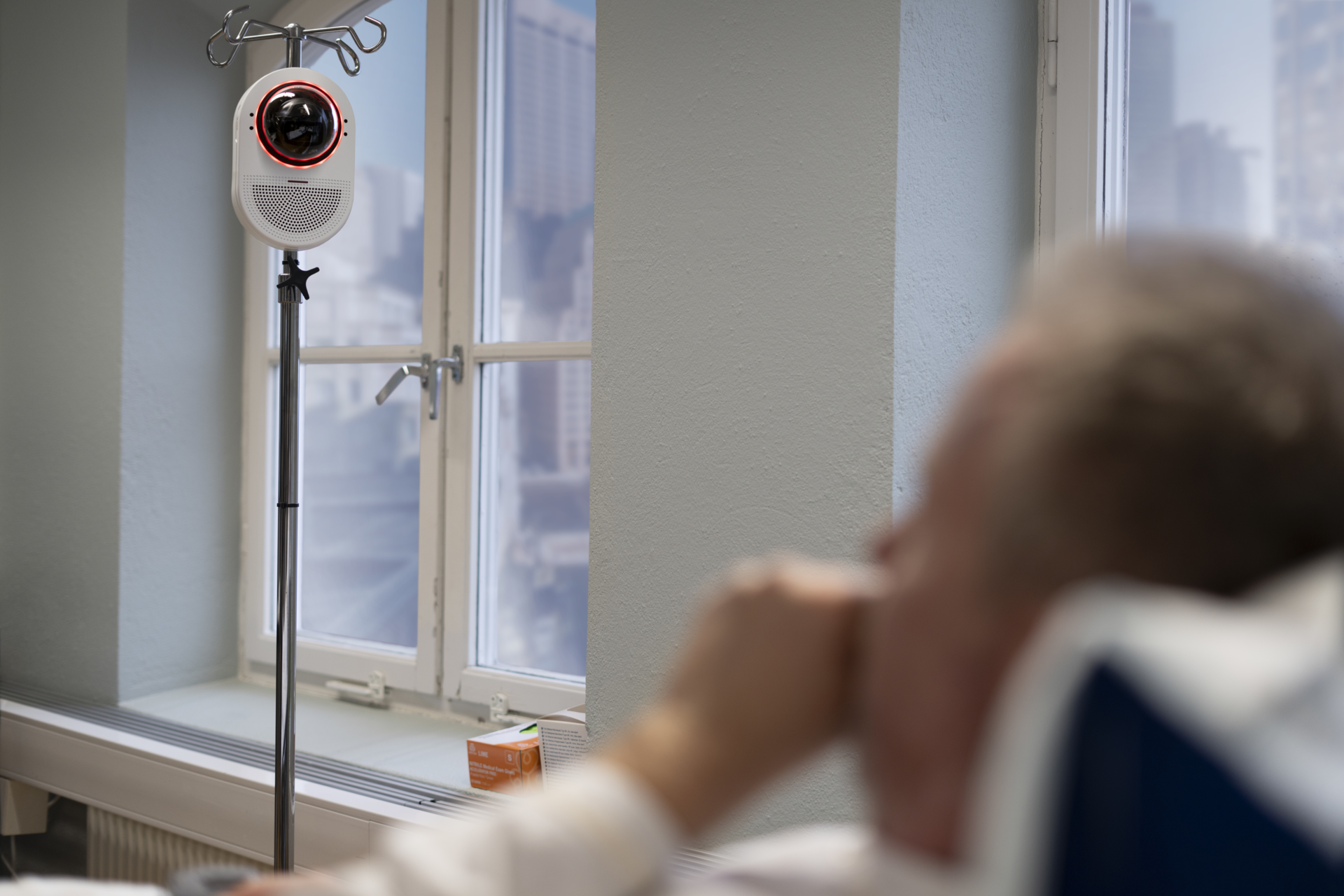 An AXIS Q9307-LV network camera mounted on a medical pole in a hospital room, facing a patient resting in bed.