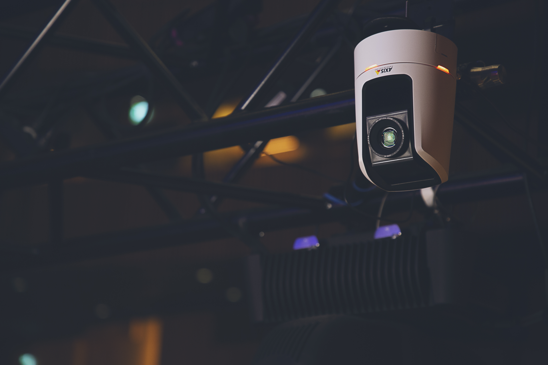 An AXIS V5938 PTZ Network Camera mounted on ceiling rigging in a darkened concert hall, with stage lighting visible in the background.