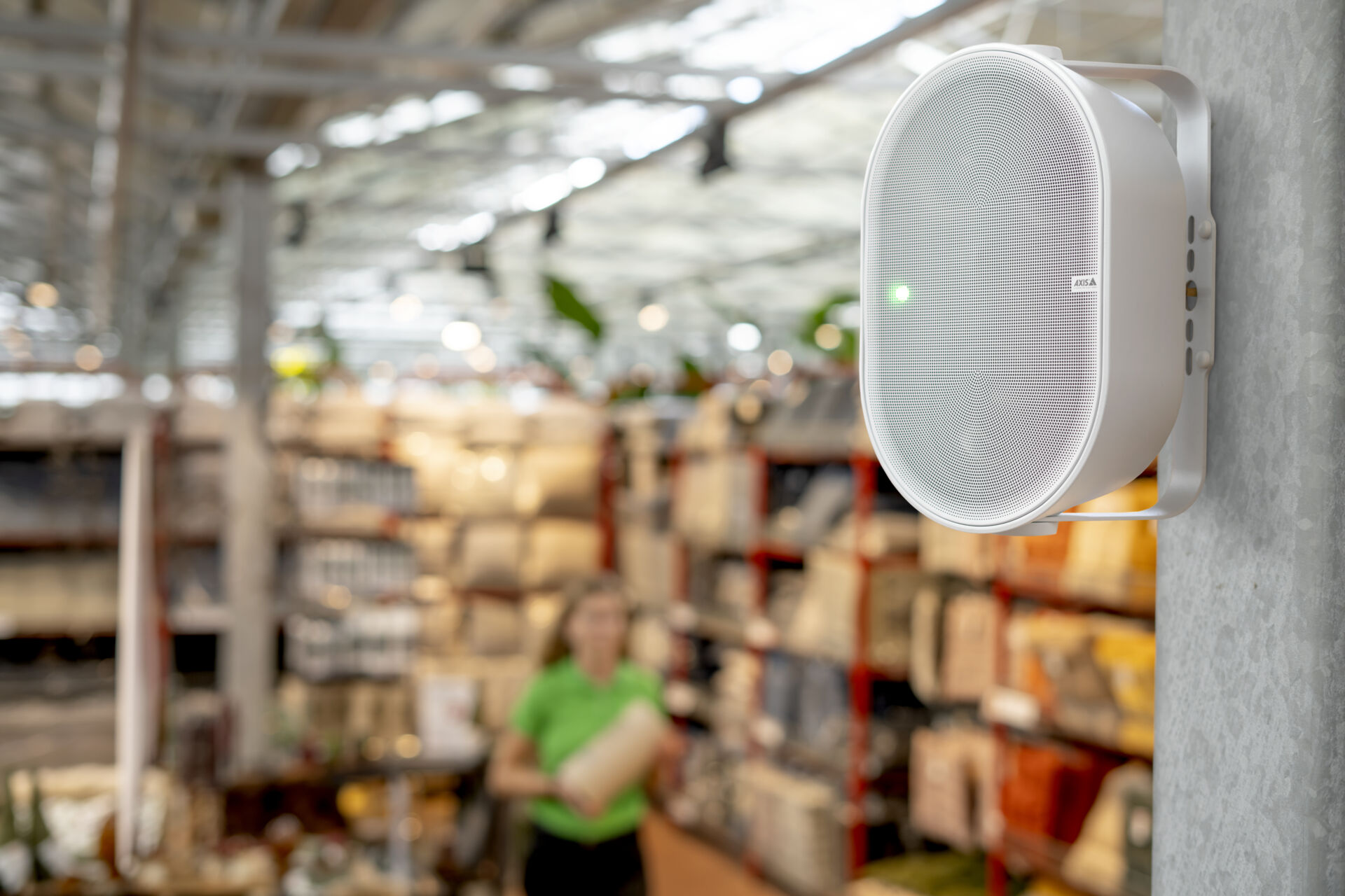 Close-up of a white, oval-shaped AXIS C1110-E network speaker mounted vertically on a concrete pillar inside a retail store. A green status light is illuminated on the speaker's mesh face. In the blurred background, a store employee wearing a bright green polo shirt walks past well-stocked shelves.