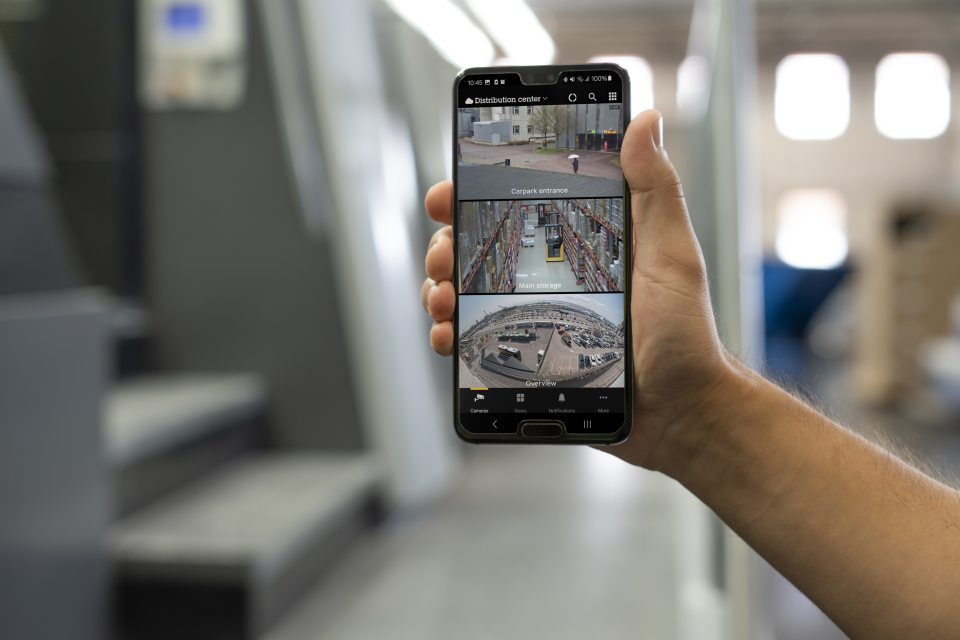 A hand holding a smartphone displaying the AXIS Camera Station Pro mobile app. The screen shows three live video feeds stacked vertically: a carpark entrance, a warehouse storage area with a forklift, and a wide-angle overview of an outdoor lot.