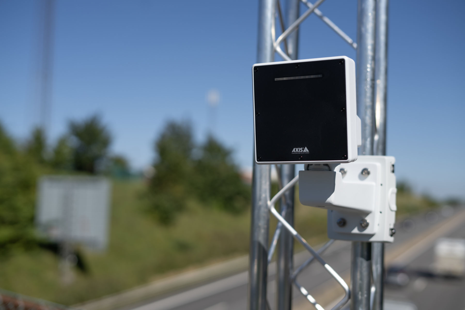 An AXIS D2210-VE network radar detector mounted on a galvanized steel truss pole alongside a highway, with trees and blue sky in the background.