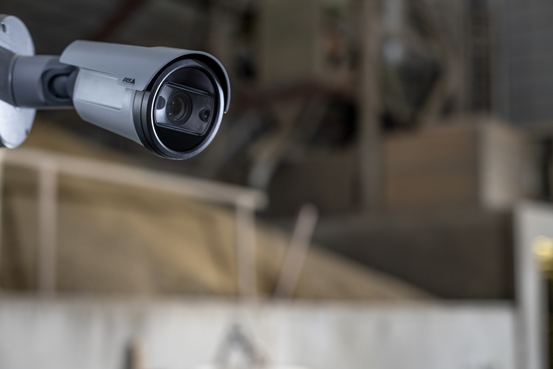 A grey AXIS P1468-XLE explosion-protected bullet camera mounted on a wall, overlooking a blurred industrial factory or warehouse environment.