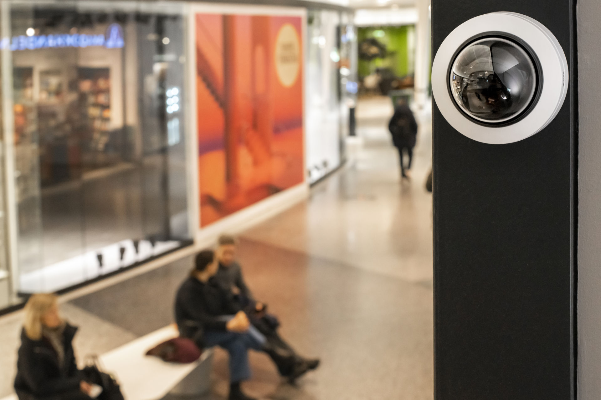 A close-up of an AXIS P32 dome camera mounted on a black pillar, with blurred shoppers in a bright mall corridor in the background.