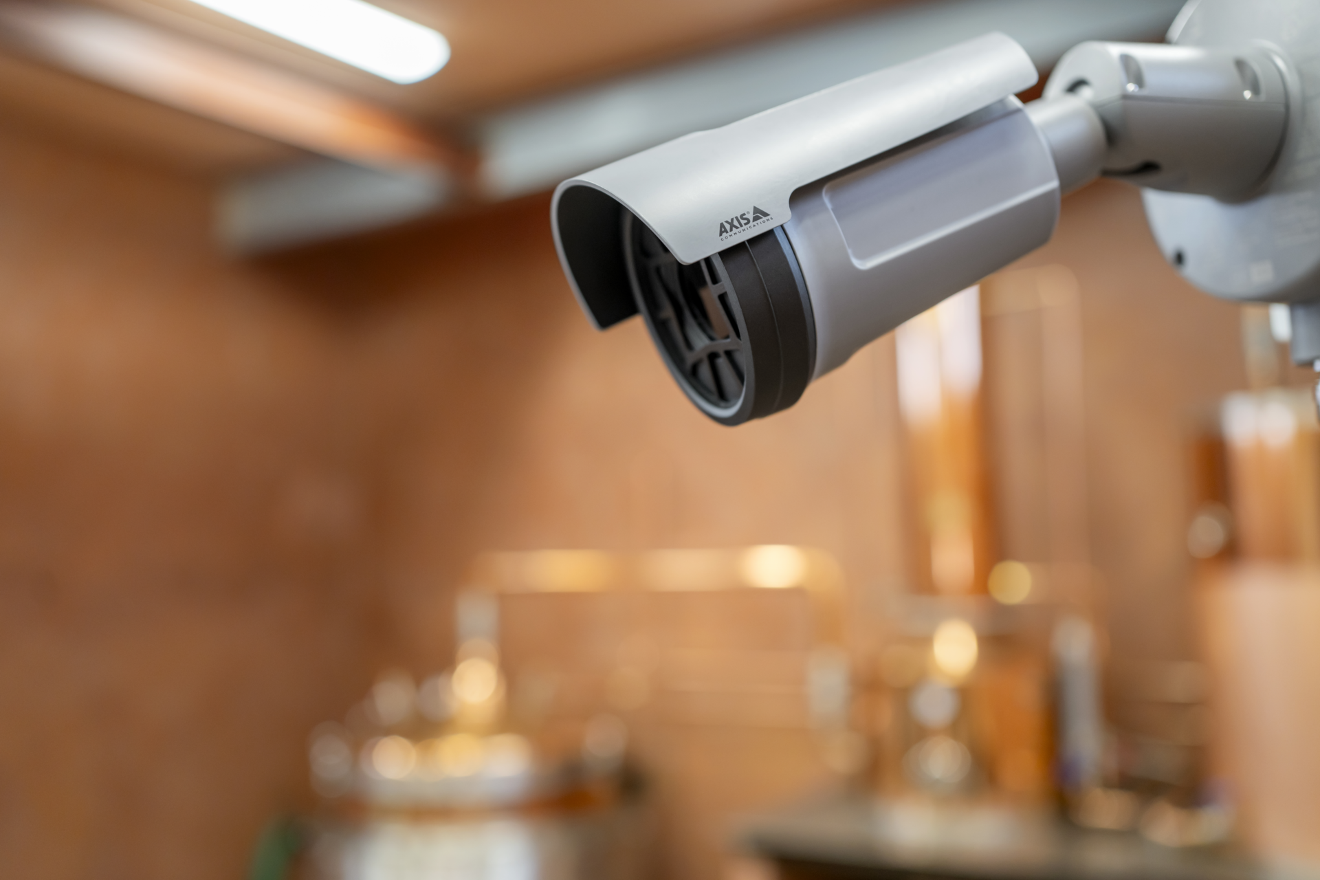 An AXIS Q1961-XTE explosion-protected thermal camera mounted on a wall bracket, overlooking blurred stainless steel brewing vats in the background.