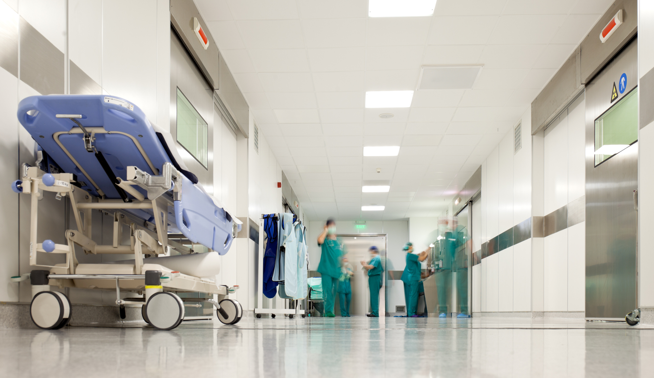 An empty blue medical stretcher parked in a brightly lit hospital hallway, with blurred medical staff in green scrubs moving in the background.