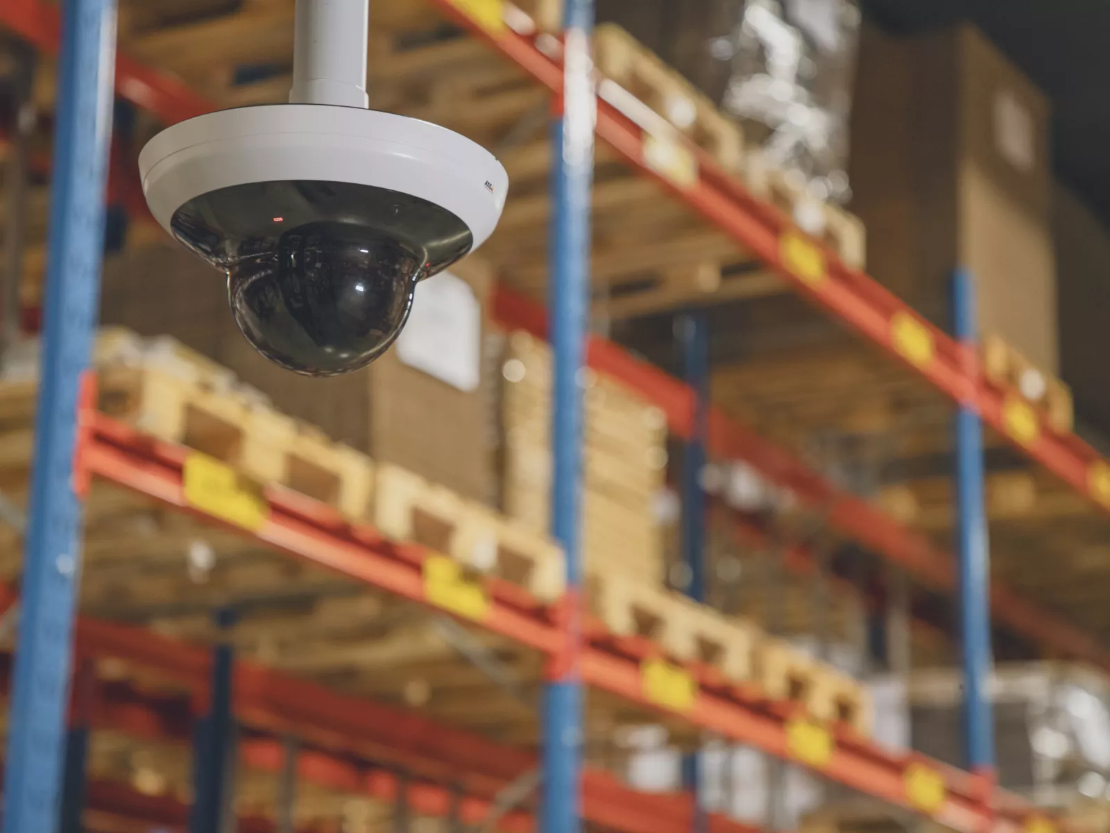 White AXIS M5000-G PTZ dome security camera mounted to the ceiling in a warehouse aisle, with blurred pallet racks in the background.