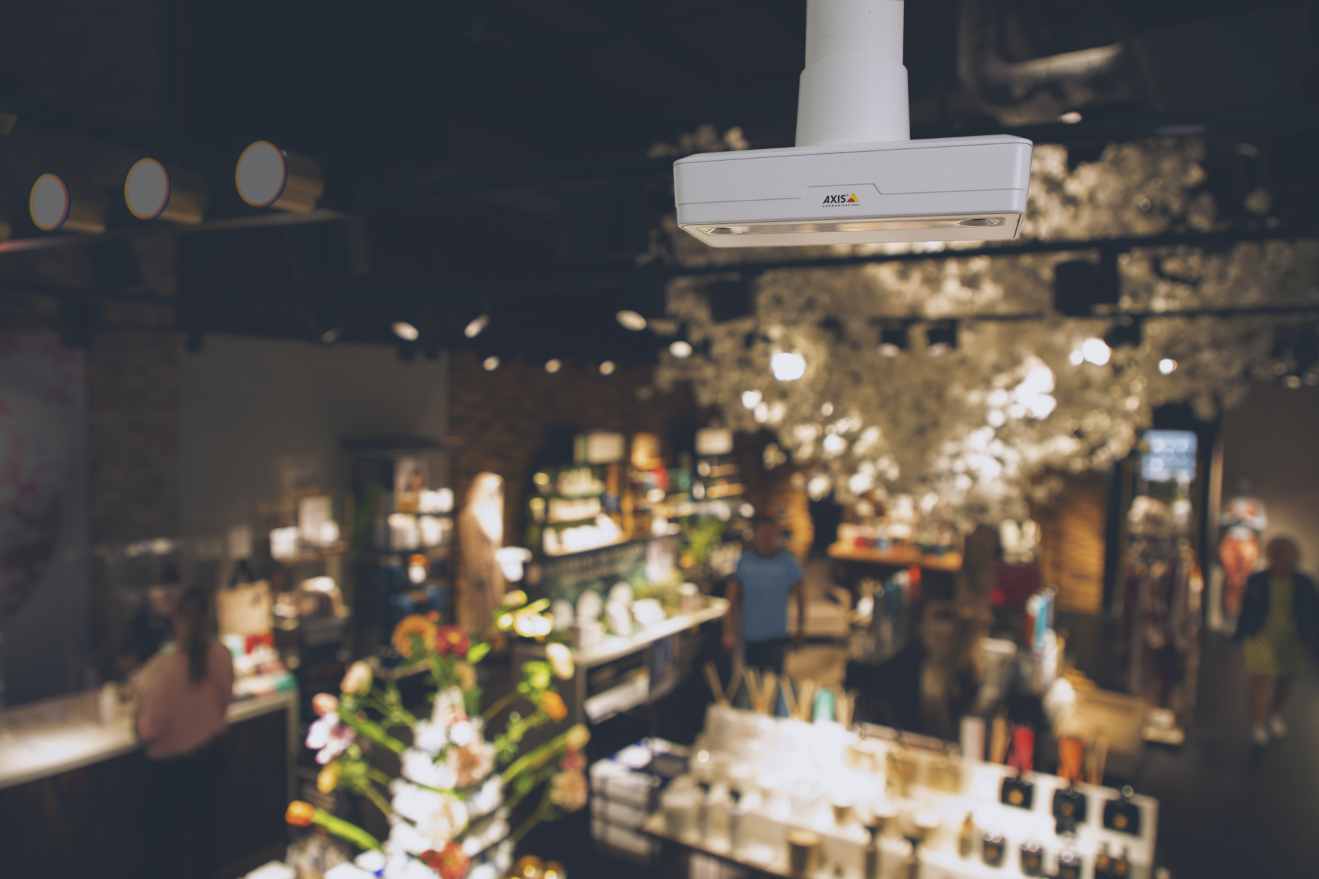 An AXIS P8815 overhead camera mounted on a white pendant from a dark store ceiling, with a blurred retail shop floor visible below featuring shoppers, display counters, warm lighting, and decorative trees.