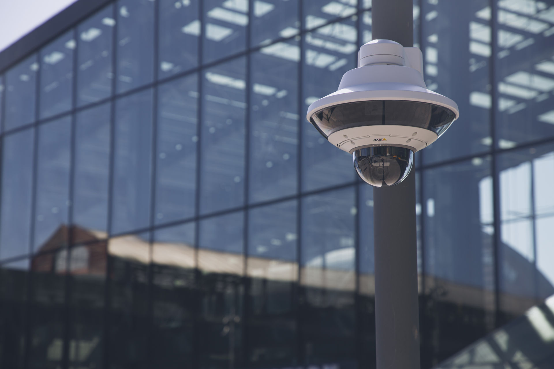 A white AXIS panoramic security camera mounted on a pole in front of a modern building with a reflective glass facade.