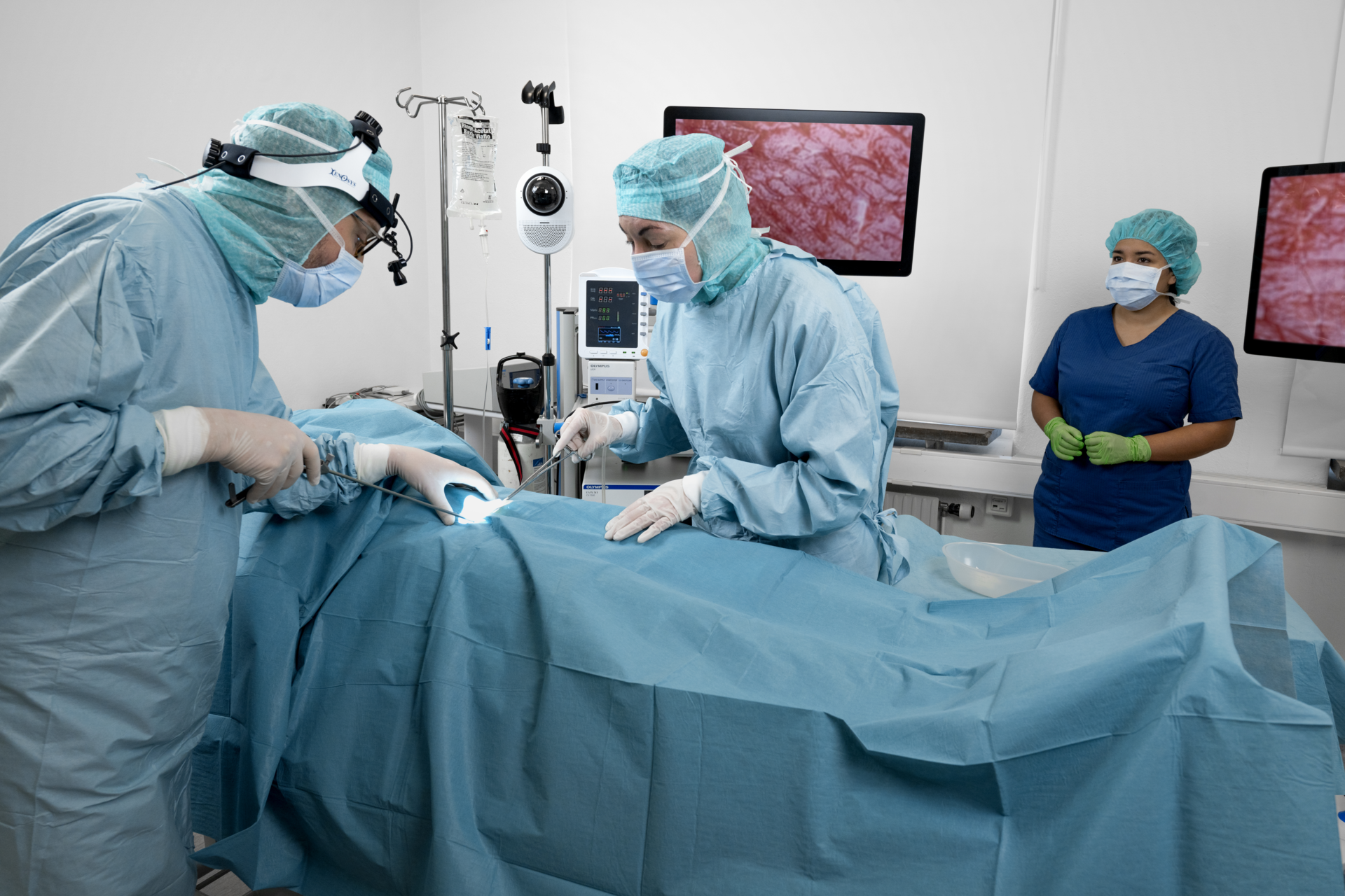 Two surgeons in blue sterile gowns and masks performing a procedure on a draped patient under a bright surgical light. A scrub nurse in blue scrubs and green gloves observes in the background. A white dome camera on a stand and two surgical monitors are positioned near the surgical table.