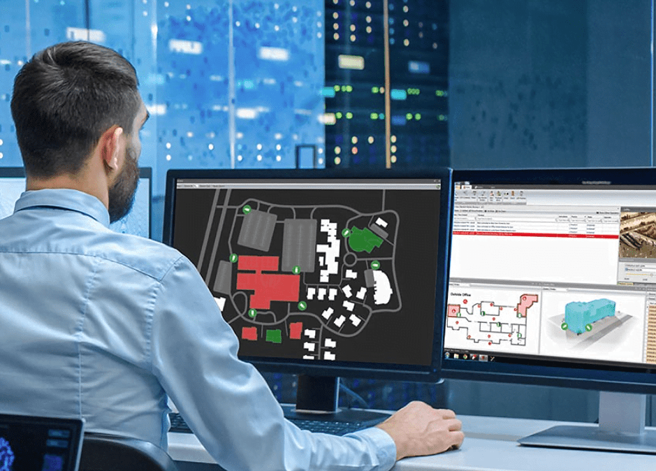 A security operator sitting at a desk in a high-tech control room, looking at dual computer monitors displaying site maps, floor plans, and system logs.