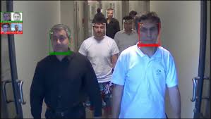 Security camera footage showing several men walking down a hallway. AI facial recognition software overlays colored bounding boxes—green, white, and red—around their faces to indicate their identification status.