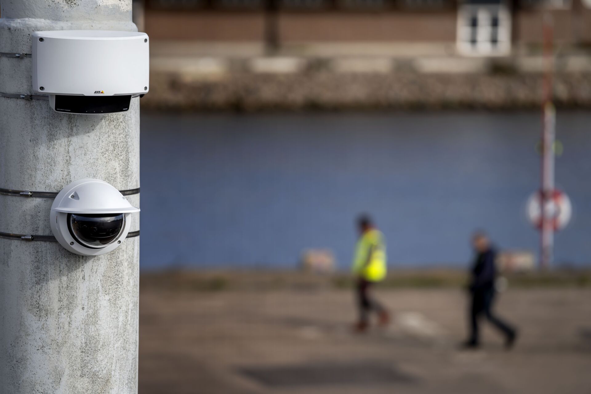 A close-up of a white AXIS security radar and a dome camera mounted on a concrete pole. In the blurred background, two people walk along a paved path next to a body of water.