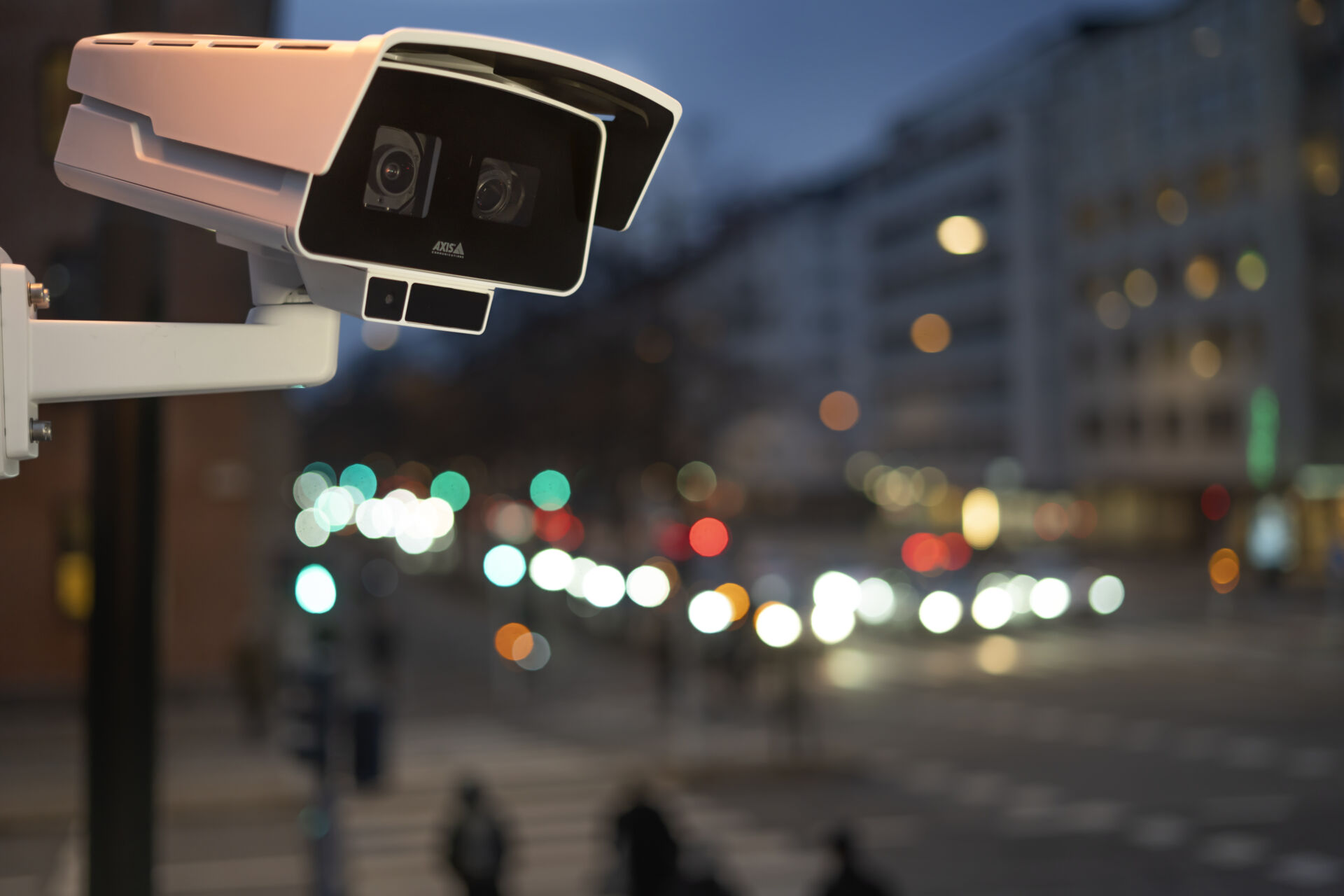 A white AXIS security camera mounted on a wall, overlooking a city street at dusk with blurred, colorful traffic lights in the background.