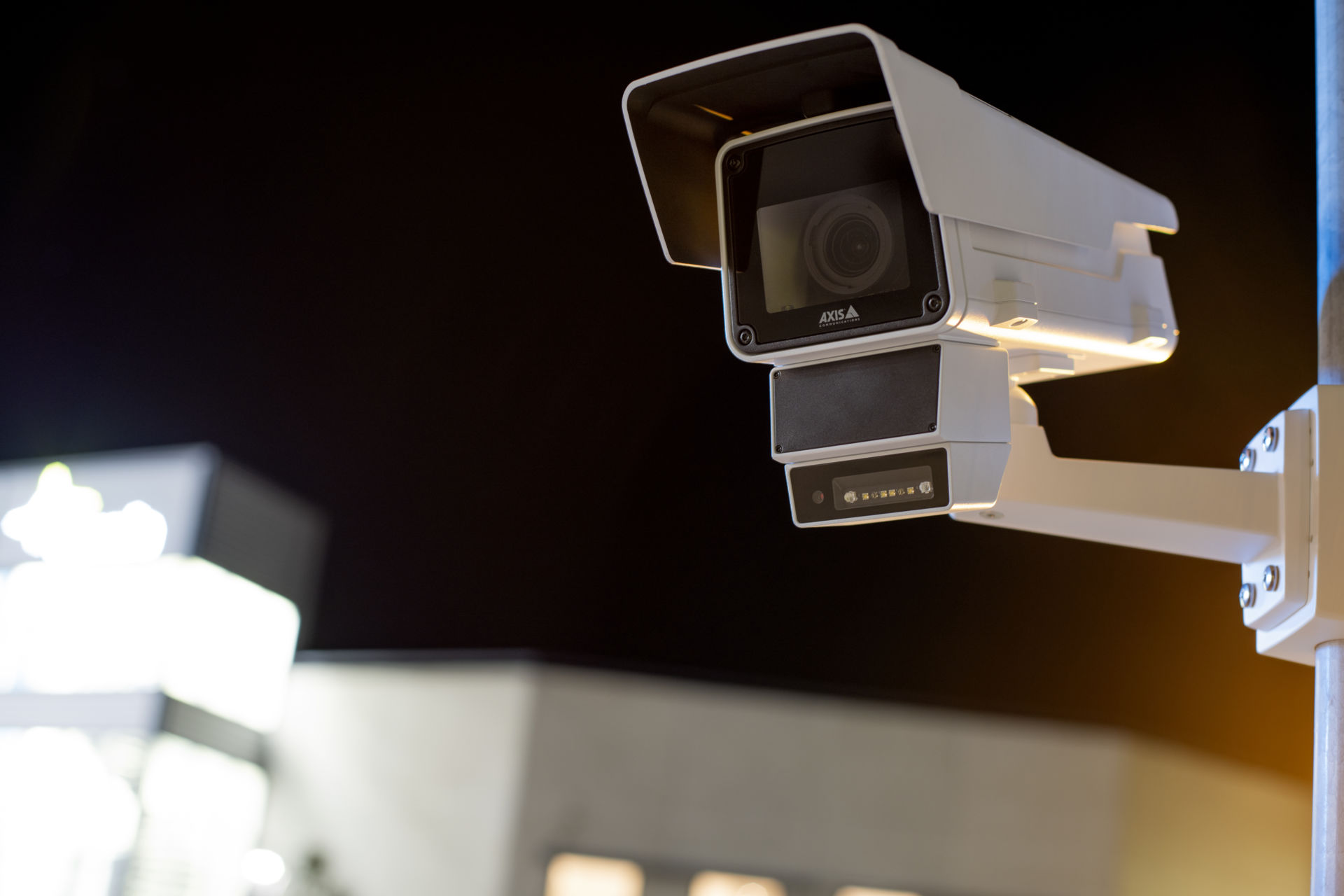 A white AXIS Communications security camera with a built-in radar and LED illuminator mounted on a pole at night, positioned against a dark sky with a blurred building in the background.